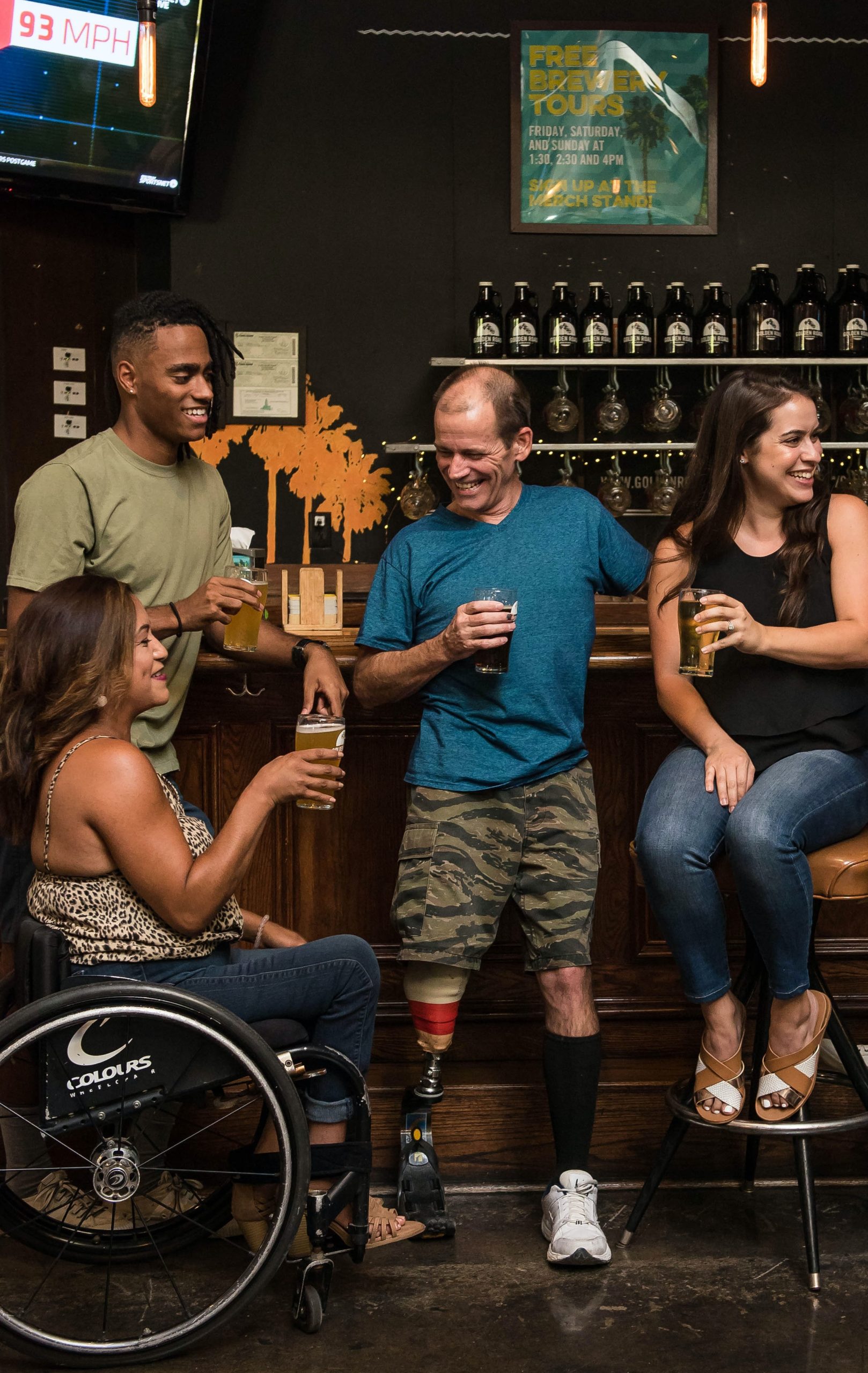 Bild von 4 Personen, zwei Frauen und zwei Männer in einer Bar, die zusammen Bier trinken wovon ein Mann eine Beinprothese hat und eine Frau im Rollstuhl sitzt